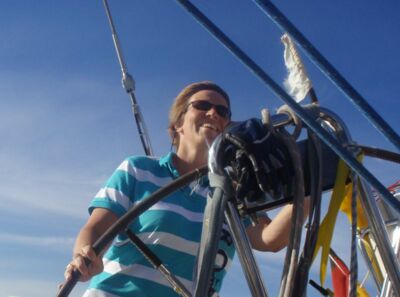 Eine Frau mit einem türkis-weiß gestreiftem Poloshirt steuert ein Segelboot